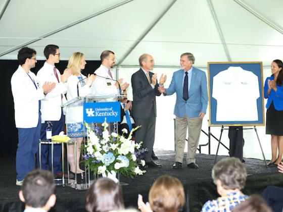 Six individuals on stage applauding while unveiling a donor's framed white lab coat, which is presented as a thank you for the donor's generosity.