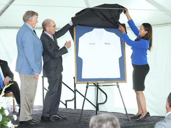 three people on stage unveiling a framed white lab coat in honor of a donor.