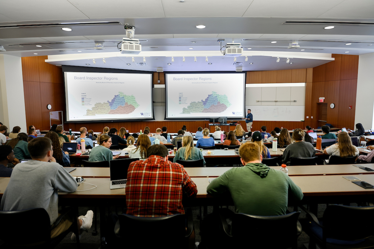 A group of students seen from behind sitting in theatre-style rows of desks with an instructor in front of a projector.