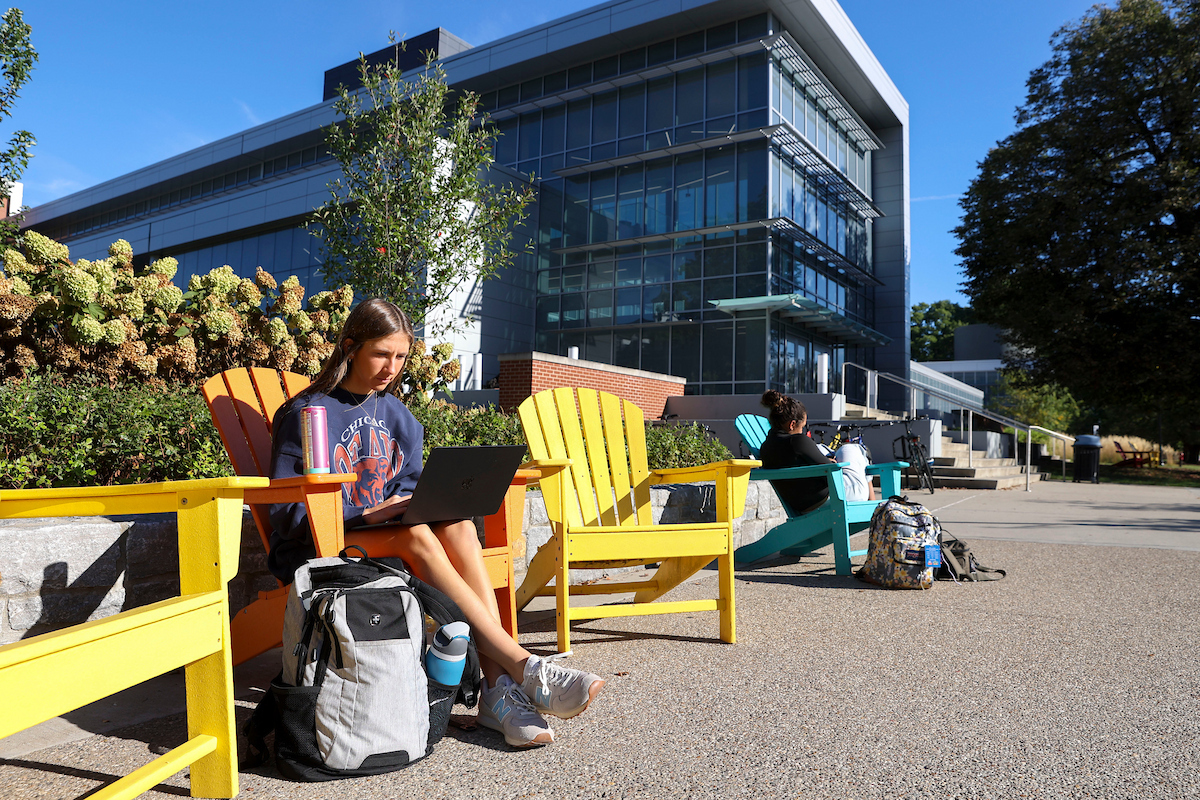 A student looks at their laptop sitting in a coloful Adirondack chair outside the Chemistry Physics Building.
