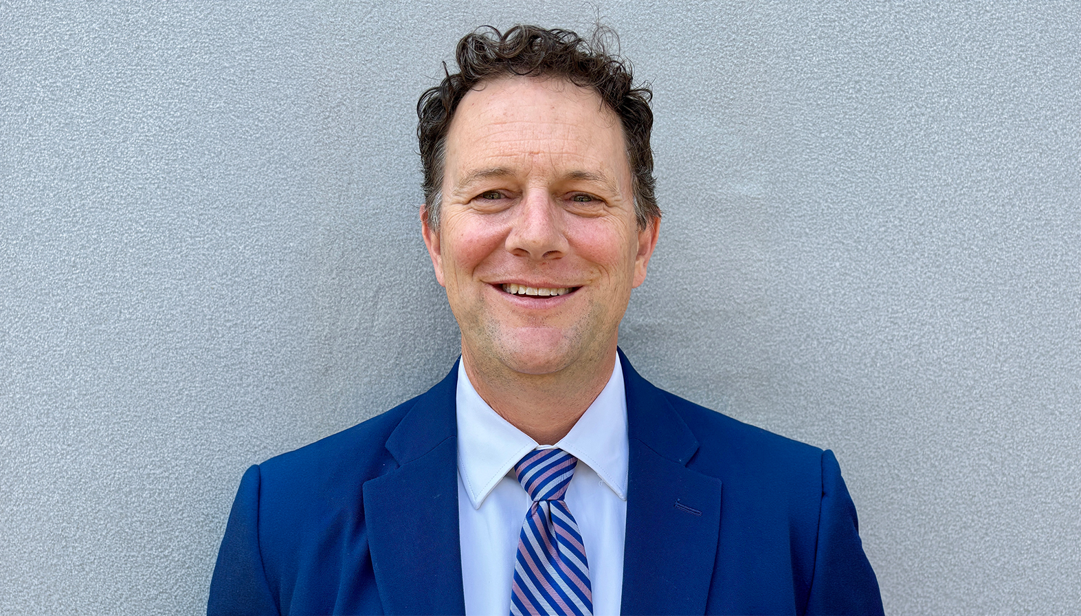 Phillip Gribble poses for a headshot, wearing a navy blue suit, white shirt and blue striped tie, against a textured gray wall.