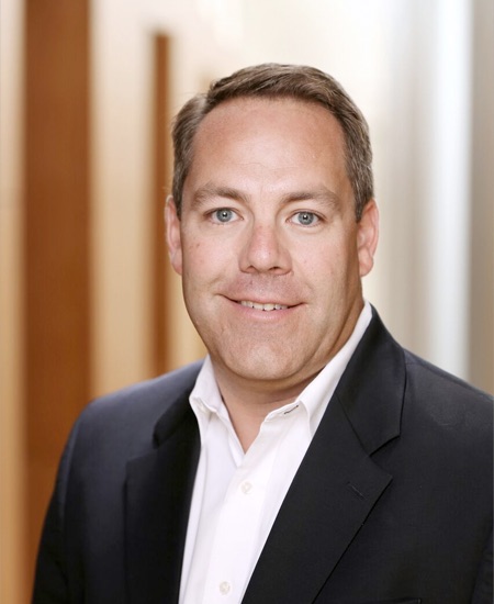 Chris Clifford headshot, wearing a black blazer and white button down shirt and a blurry wood and white background. 
