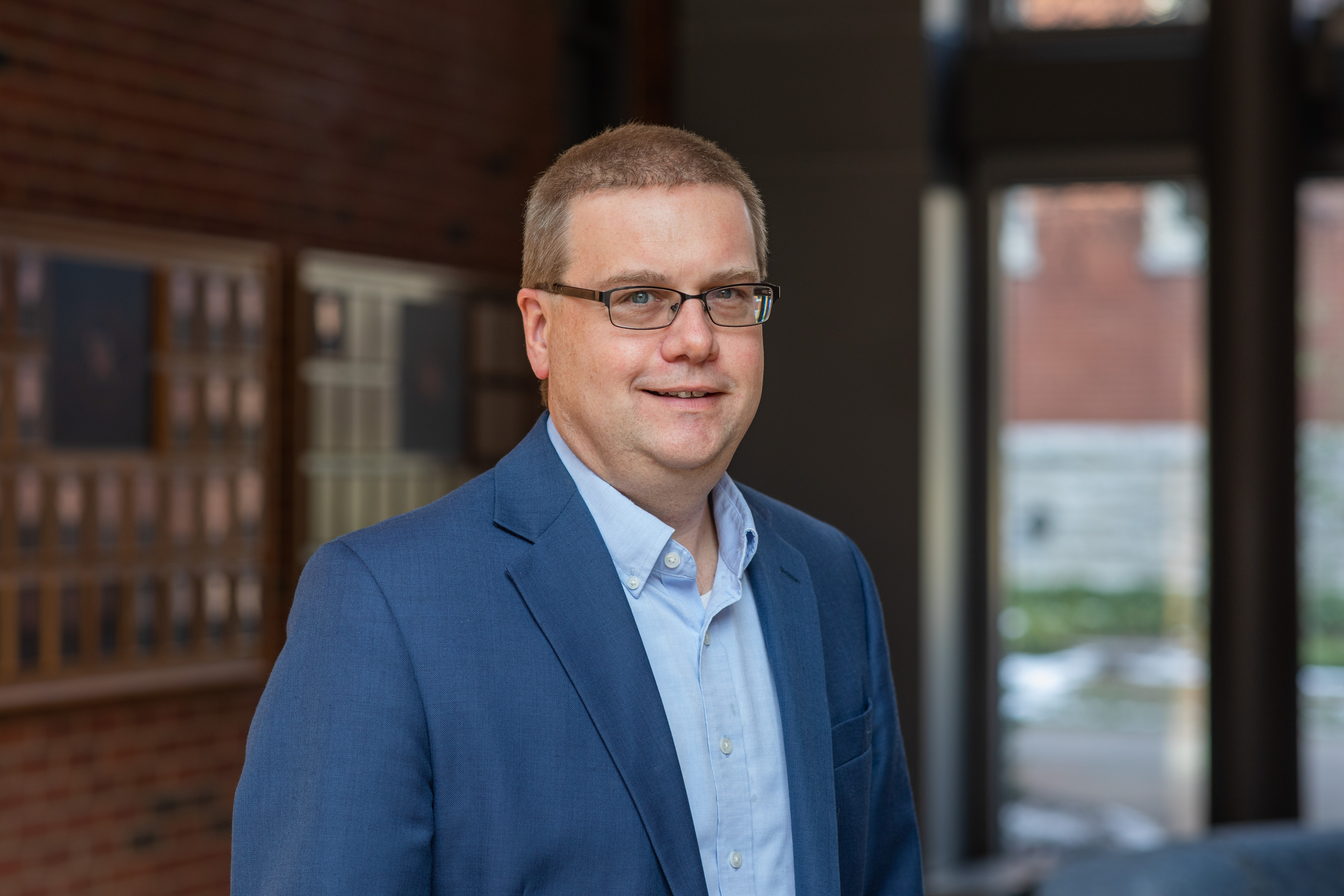 headshot of Mike Renfro, wearing a light blue blazer and button down, inside a brick building.