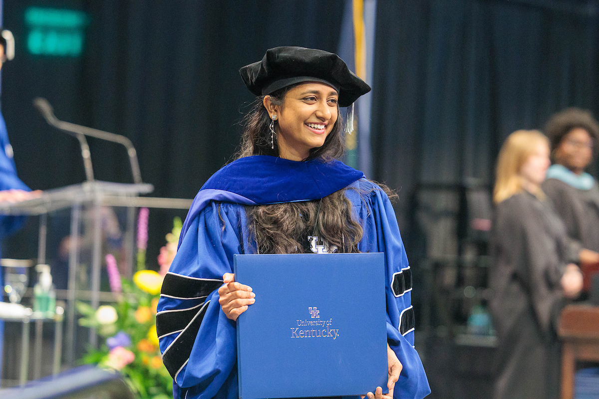 A UK doctoral student wearing blue graduation regalia holds their diploma and poses for a photo on the commencement stage.
