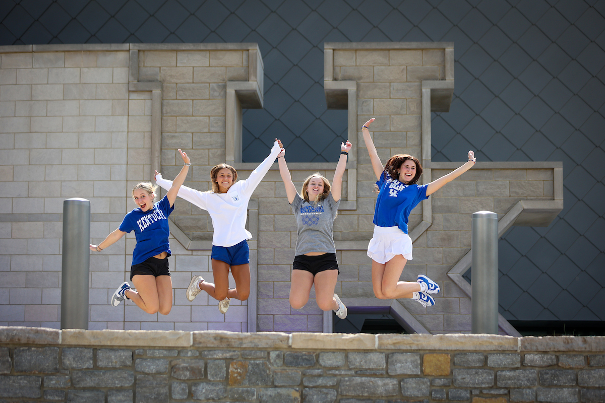 Four students jump in the air and pose in front of the large UK stone sign with a low stone wall in the background. 