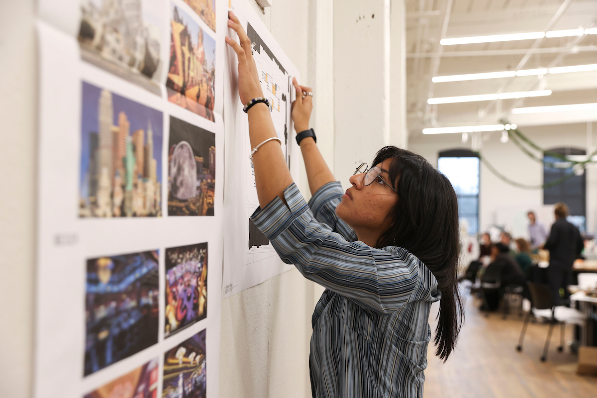 A student in a plaid shirt presses their hands against a poster to ensure it sticks to a wall, which is filled with other photos of designs and images.