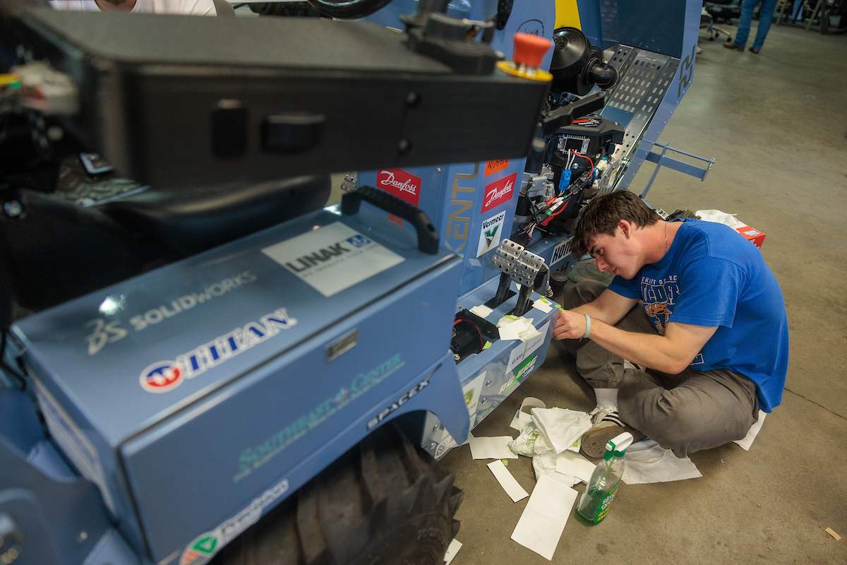 A student wearing a blue shirt sits on the ground next to a large tractor, affixing something to its side.