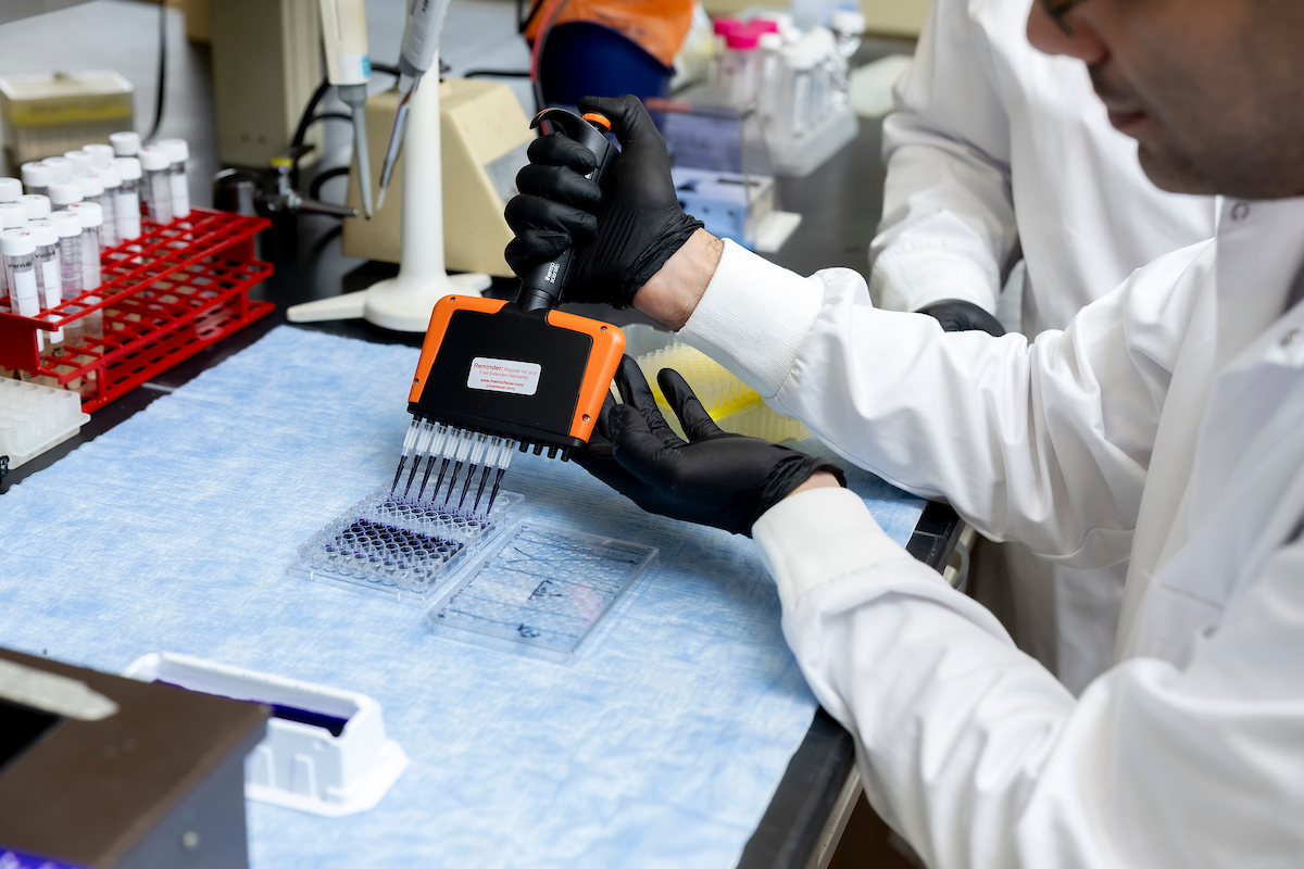 A person wearing a white lab coat uses a pipette to inject black liquid into a plastic container while at a lab bench filled with test tubes and equipment.