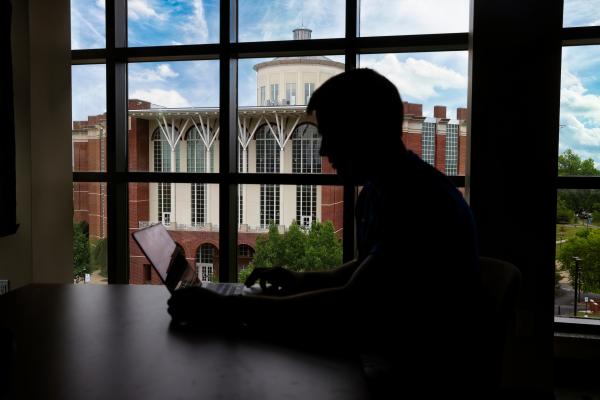 A view out of a top floor window in Lewis Hall where the William T. Young Library is seen in front of a partly cloudy, blue sky. There is a silhouette of someone sitting by the window looking at a laptop.