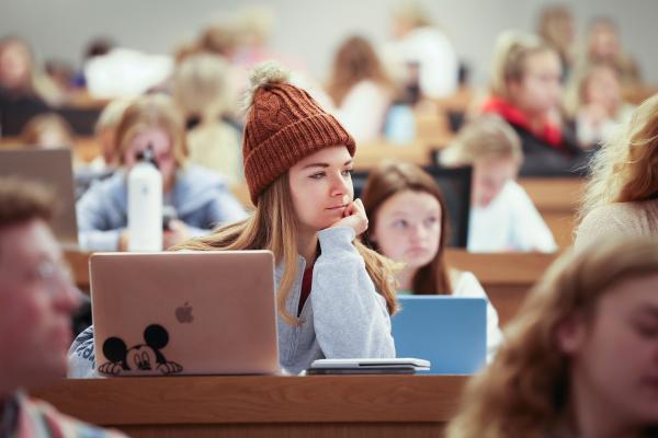 a student wearing a reddish-brown beanie sits inside a lecture hall with a laptop and their head propped on their hand. In the background, there are tiered rows of other students sitting.