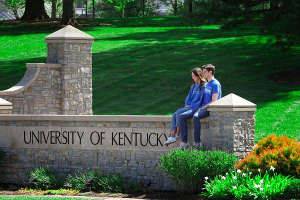 Two students in blue UK shirts sitting on a stone sign that says "University of Kentucky"