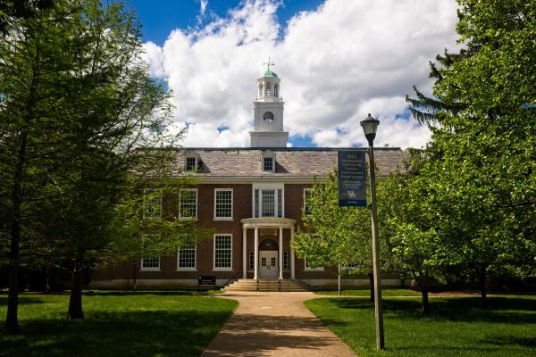 Two-story brick building with a tall cupola centered against a bright blue sky and white clouds.