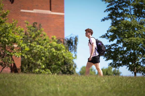 Young man in profile in a white tee and black shorts with a black backpack walking with grass in the foreground and trees and a brick building set against a pale blue sky in the background.