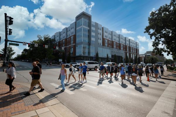 People crossing the street with a large, five-story building in the background and a blue sky with clouds.