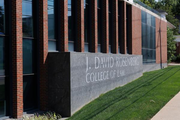 Side-along view of a gray stone sign that says "J. David Rosenberg College of Law" set in front of a building with red brick and glass facade.