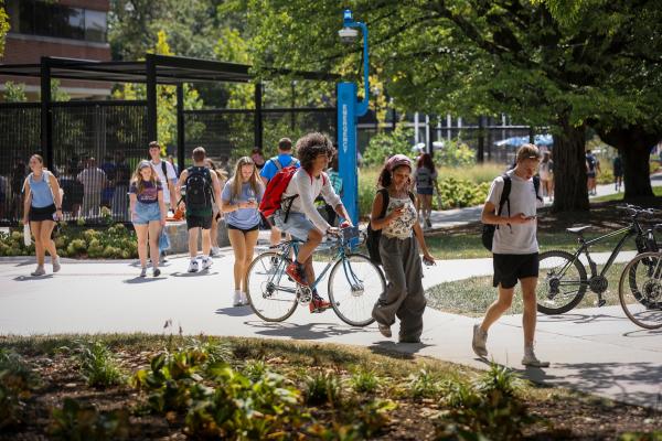 Students walking and biking along a sidewalk with flowers and trees in the background.