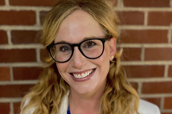 Blonde woman wearing royal blue shirt and white blazer with glasses in front of red brick wall