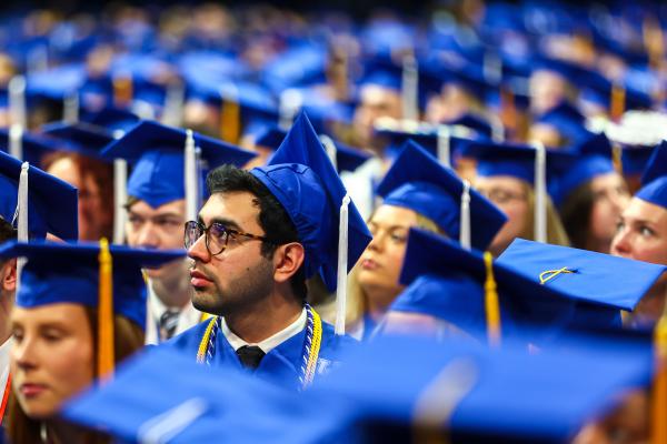 an individual wearing glasses and a UK blue graduation cap and gown looking to the side, surrounded by other, out-of-focus people wearing the same caps and gowns