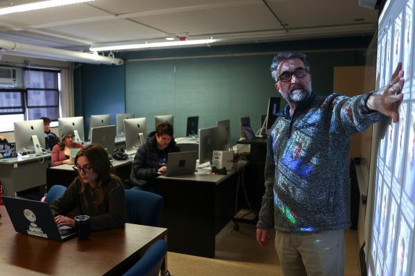 dim computer lab with students looking at computers and instructor at the front gesturing at a projector screen.