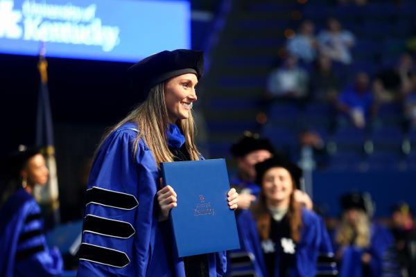 A UK doctoral graduate wearing regalia posing for a photo on stage holding their diploma.