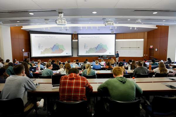 A group of students seen from behind sitting in theatre-style rows of desks with an instructor in front of a projector.