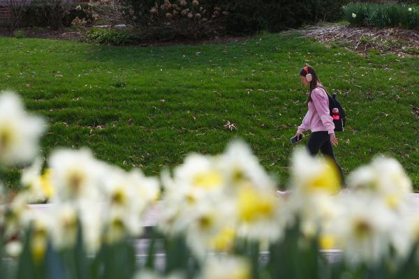 A student walking across the background with headphones and a pink sweater in springtime weather.