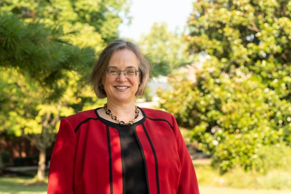 Rebecca Thomas, wearing a red blazer, posed in front of a sunny tree-lined field.