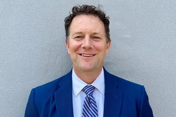 Phillip Gribble poses for a headshot, wearing a navy blue suit, white shirt and blue striped tie, against a textured gray wall.