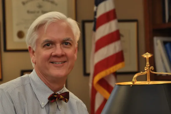 Greg Van Tatenhove wearing a light blue button up shirt and a red bowtie inside an office with a lamp and U.S. flag.