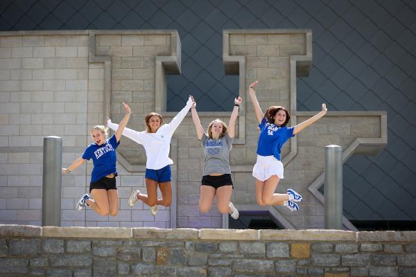 Four students jump in the air and pose in front of the large UK stone sign with a low stone wall in the background. 