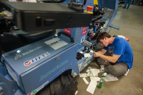 A student wearing a blue shirt sits on the ground next to a large tractor, affixing something to its side.