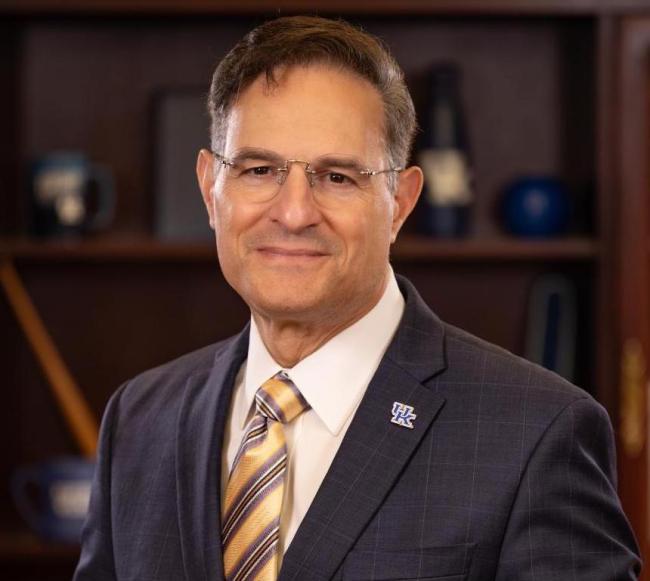 Headshot of man wearing glasses and navy blazer, white button-up and gold striped tie.