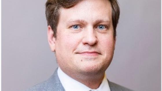 Headshot of male with short, brown hair wearing a gray suit with a blue tie and white button-up shirt. 