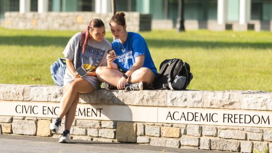 two students sitting on a low stone wall, looking at one of their phones. It is a sunny day, and there is a small field of green grass behind them. 
