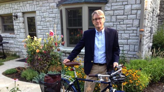 Ned Crankshaw standing with a bike and backpack in the bike's basket in front of a stone-faced house. 