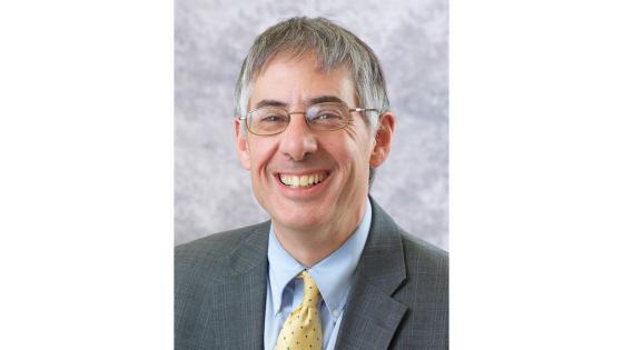Paul E. Salamanca wearing a gray suit and light blue shirt with a yellow tie, posed in front of a gray background. 