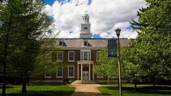 Two-story brick building with a tall cupola centered against a bright blue sky and white clouds.