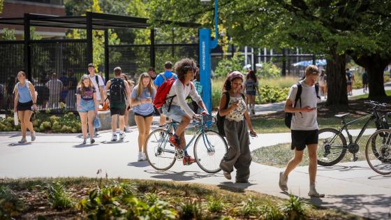 Students walking and biking along a sidewalk with flowers and trees in the background.
