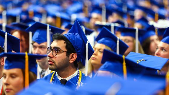 an individual wearing glasses and a UK blue graduation cap and gown looking to the side, surrounded by other, out-of-focus people wearing the same caps and gowns
