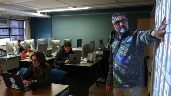 dim computer lab with students looking at computers and instructor at the front gesturing at a projector screen.