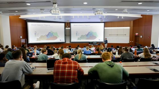 A group of students seen from behind sitting in theatre-style rows of desks with an instructor in front of a projector.