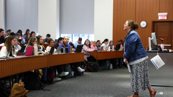 A classroom with an instructor standing in front of a rows of students seated at desks.