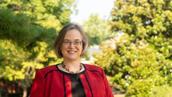 Rebecca Thomas, wearing a red blazer, posed in front of a sunny tree-lined field.