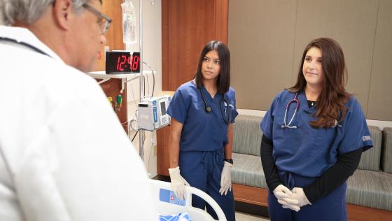 Two nursing students in scrubs listen to an instructor during a simulation exercise.