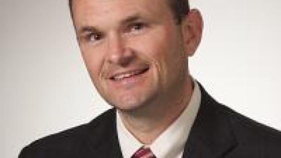 Headshot of Matt Bacon in a suit and red striped tie.