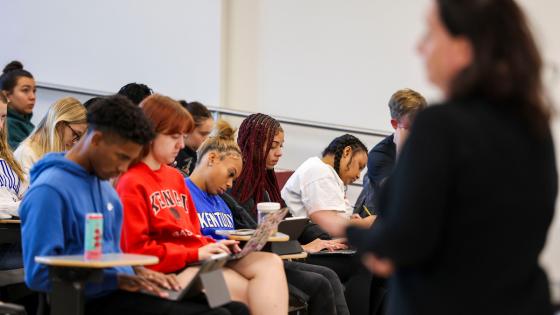 An instructor stands in front of a class full of seated students. 
