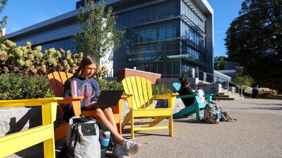 A student looks at their laptop sitting in a coloful Adirondack chair outside the Chemistry Physics Building.
