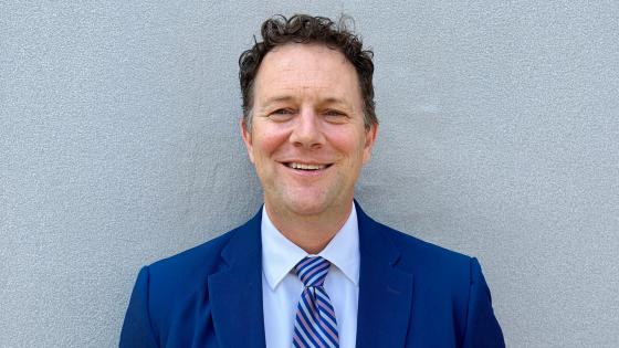 Phillip Gribble poses for a headshot, wearing a navy blue suit, white shirt and blue striped tie, against a textured gray wall.