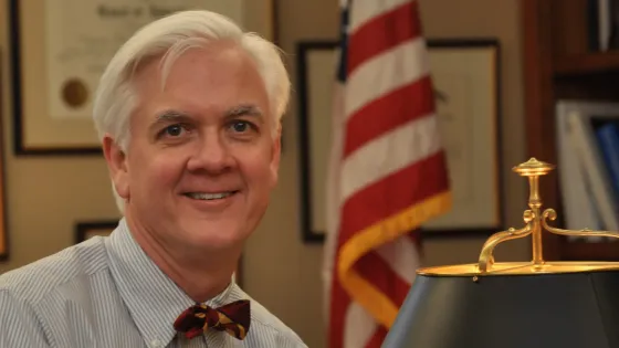 Greg Van Tatenhove wearing a light blue button up shirt and a red bowtie inside an office with a lamp and U.S. flag.