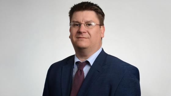 James Buss, wearing a navy blue suit with maroon tie and light blue button down shirt, poses for a headshot against a light gray background.